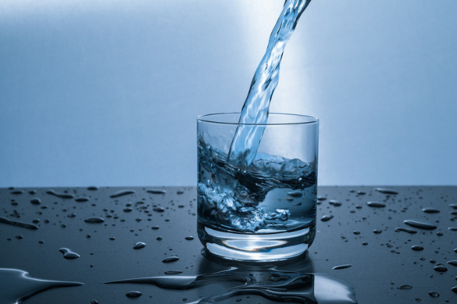 Water being poured from a height at an angle into a glass. The image is aqua blue with droplets of water all around the glass.